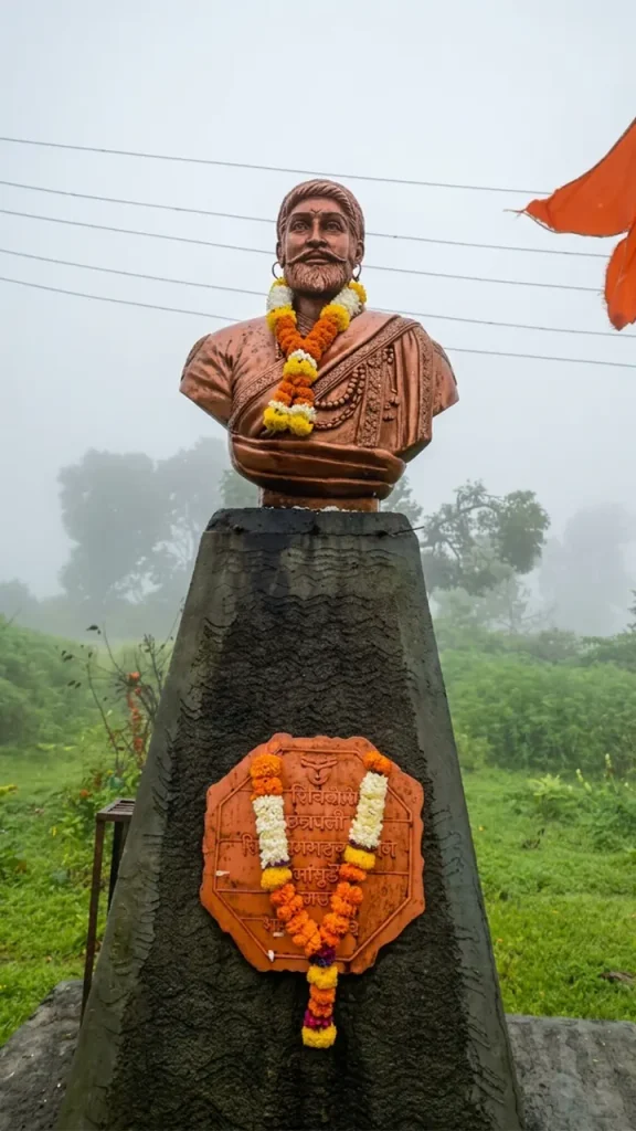 Bronze bust of Chhatrapati Shivaji Maharaj adorned with flower garlands in a misty landscape with a saffron flag