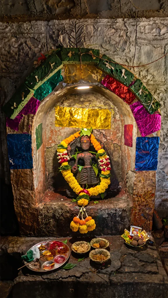 Ancient carved stone idol of a deity placed inside a recessed stone wall niche, decorated with colorful metallic foil, fresh marigold garlands, and ritual puja offerings on small plates below